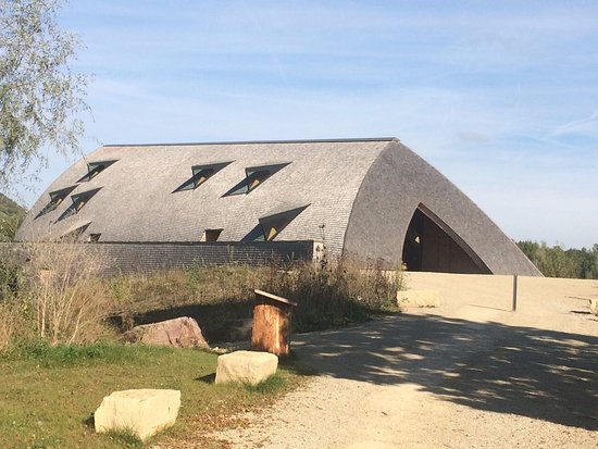 Curved brick building with a large arched entrance and small triangular windows, set in a sunny park.